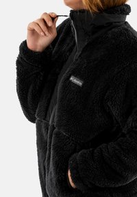 Chaqueta de forro polar negra con cierre frontal, cuello alto y detalles de tela negra en contraste. Presenta una textura acogedora y dos bolsillos laterales.