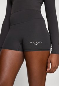 Fekete atletikai rövidnadrág rugalmas anyagból, sima felülettel és síköltéses varrásokkal. Az elején "HYROX" felirat és egy fehér logó található.