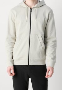 Felpa grigia chiara con zip, tasca frontale, cappuccio con cordino e zip nera a contrasto. Presenta una toppa tonale con logo sul petto.