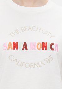 Valge puuvillane T-särk, millel on masinõmmeldud tekst. "RANDLINN" ja "CALIFORNIA '95" beežis, "SANTA MONICA" värvikates pastelltoonides.