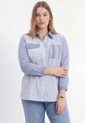 Woman wearing a blue and white striped button-up shirt with two chest pockets, blue jeans, and gold rings and bracelet, standing with arms crossed.