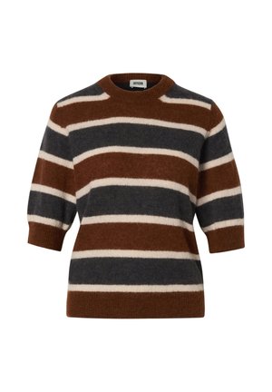 Jeune femme portant un pull rayé marron, noir et beige, un jean large bleu clair et des chaussures noires, debout devant un fond uni.