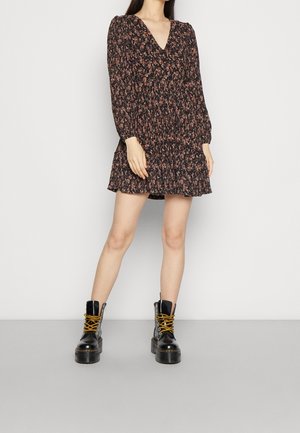 Femme portant une robe courte noire à fleurs avec des manches longues, associée à des bottes noires épaisses à lacets jaunes.