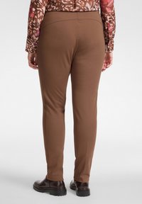 Brązowe dopasowane spodnie o gładkiej fakturze, proste nogawki i średni stan, zestawione z ciemnymi butami. Tło jest neutralne.