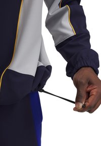 La chaqueta deportiva de tela en azul marino y gris presenta puños elásticos, un cordón en el dobladillo y detalles en amarillo contrastante.