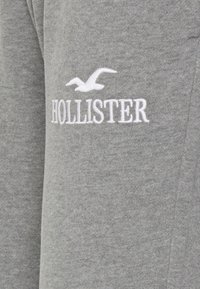 Grått tyg med vit broderad Hollister-logotyp som visar en flygande mås ovanför varumärket.