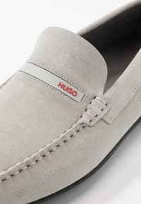 Mocassins en daim gris dotés d'un design cousu et d'une semelle en caoutchouc lisse. Une bande en caoutchouc avec le logo "HUGO" en rouge souligne le dessus.