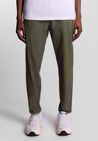 Olijfgroene sportbroek, gemaakt van lichtgewicht materiaal, heeft een slim fit ontwerp met een logoaccent nabij de taille. Gepaarde met witte sneakers.