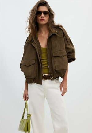 Kobieta nosząca oversize brązową kurtkę, oliwkowozielony top, białe spodnie, okulary przeciwsłoneczne, trzymająca małą zieloną torebkę na jednolitym jasnym tle.