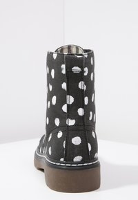 Bottine en tissu noir à pois blancs, semelle plate marron et coutures blanches contrastantes. Dispose d'une languette à l'arrière.