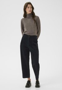 Bruin coltrui gecombineerd met donkerblauwe corduroy broek met een wijd model, voorzakken en een knoopsluiting. Zwarte loafers maken de look af.