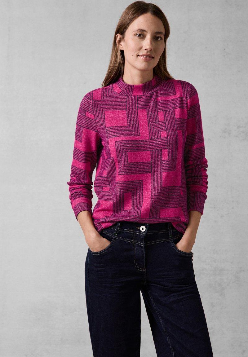 Fuchsia trui met geometrisch zwart patroon, hoge halslijn en geribbelde manchetten, gecombineerd met donkerdenim broek met voorzakken.