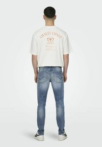 Witte katoen T-shirt met "AMALFI COAST" opdruk in perzik, gedragen met lichtblauwe skinny jeans en zwart-accentueerde witte sneakers.