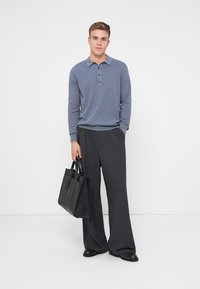 Graues langärmliges Polohemd mit drei Knöpfen, kombiniert mit lockeren schwarzen Hosen und einer großen schwarzen Ledertasche. Schlichter Hintergrund.