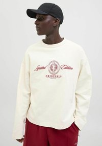 Sudadera color crema con mangas largas, que presenta texto y emblema en burdeos. Combinada con pantalones rojo oscuro y una gorra negra.