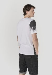 T-shirt blanc avec un motif d'éclaboussures de peinture noire sur les épaules, associé à un short cargo foncé. Matière en coton, coupe décontractée, manches courtes.