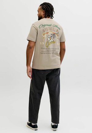 Uomo con capelli intrecciati che indossa una t-shirt beige con grafica floreale e testo, jeans neri e sneaker nere, di spalle.