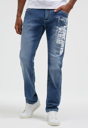 Mann trägt blaue Jeans mit weißem grafischem Text am rechten Oberschenkel und weißen Turnschuhen, steht vor einem einfarbigen Hintergrund.