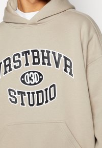 Beige hoodie met een kangoeroezak, voorzien van dikke zwarte letters: "WRSTBHVR 03D STUDIO." Zachte textuur, ontspannen pasvorm en verstelbare capuchon.