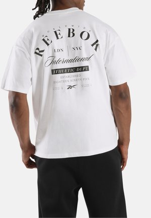 Mann trägt weißes Reebok T-Shirt mit schwarzem Text und Logos auf dem Rücken, kombiniert mit schwarzen Hosen, von hinten gezeigt.