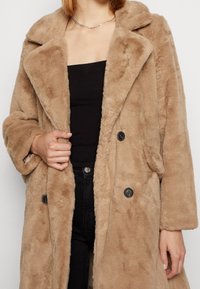 Brązowy płaszcz z imitacji futra z szerokim kołnierzem, dwurzędowym zapięciem i dużymi czarnymi guzikami. O długości do połowy uda, miękka, teksturowana powierzchnia.