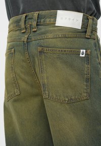 Pantaloni din denim de culoare verde măslin, cu talie înaltă, cusături vizibile, buzunare la spate și o etichetă albă pe talie, cu text în limba japoneză.