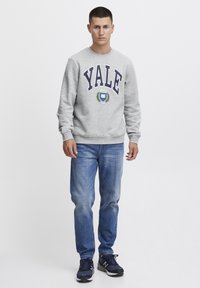 Sudadera gris con "YALE" en azul marino, con un logo circular y acentos de laurel. Combinada con jeans de un azul claro y zapatillas azul marino.