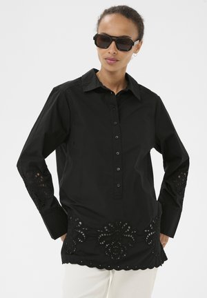 Kobieta w czarnej bluzce z guzikami i kwiatowym haftem z wycięciami oraz w dużych czarnych okularach przeciwsłonecznych, stojąca na jednolitym tle.