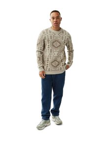 Mann trägt einen beigen Pullover mit geometrischem Muster, blaue Jeans und graue Sneakers und steht mit einer Hand in der Tasche.