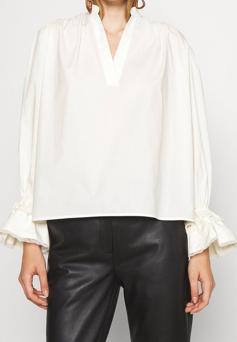 Femme portant un chemisier blanc à manches longues avec des poignets à volants et un pantalon en cuir noir sur un fond blanc uni.