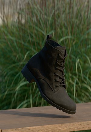 Botte en cuir marron foncé à lacets flottant au-dessus d'une surface en bois avec de l'herbe verte haute floue en arrière-plan.
