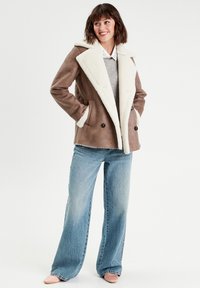 Brauner, synthetischer Shearling-Mantel mit doppelter Knopfleiste, breitem weißem Kragen, zwei Fronttaschen und kombiniert mit hellblauen, weit geschnittenen Jeans.