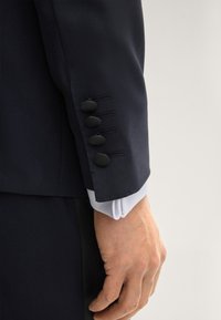 Manica di giacca da completo blu navy con quattro bottoni neri, vestibilità su misura, tessuto liscio e polsino di camicia bianco che spunta sotto.