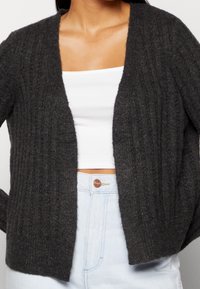 Cardigan a coste grigio scuro con scollo a V, apertura frontale e texture morbida. Abbinato a una maglietta bianca e jeans azzurri chiari.