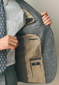 Hallime tekstureeritud hall ülikond, mis on kaetud beeži satiinvoodriga, sisepocketid ja etikett. Seljas on heledate triipudega särk ja mustriga lips.