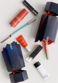 Varie cosmetiche su uno sfondo chiaro: un blush stick rosso, lucidalabbra e creme in tubetti e una scatola con accenti blu scuro e oro.