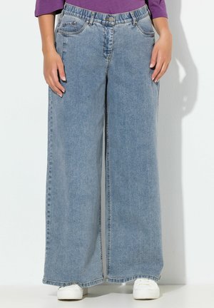 Kvinde iført lyseblå jeans med vide ben, hvide sneakers og en lilla langærmet top, stående op ad en hvid væg.