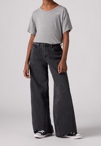 T-shirt en coton gris à manches courtes avec un petit logo, associé à un jean noir en denim à jambes larges et des baskets noires.