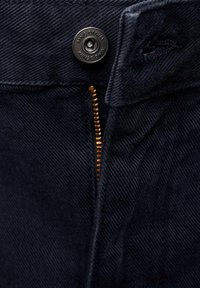 Gros plan sur un tissu en denim foncé avec un bouton en métal portant l'inscription "MNG DENIM" et une fermeture éclair en laiton partiellement visible.