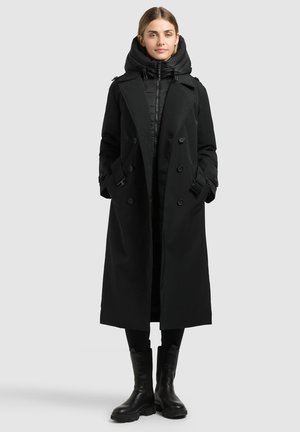 Femme portant un long manteau noir croisé sur une veste matelassée noire à capuche, pantalon noir et bottes noires, debout devant un fond uni.