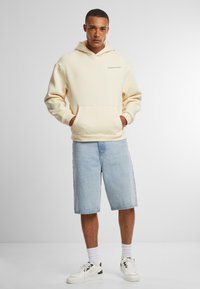 Cremefarvet hættepullover med en frontlomme, parret med lyseblå denimshorts. Tilbehør inkluderer hvide sneakers og hvide sokker.
