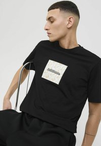 Sort bomulds T-shirt med et firkantet lap med et cremefarvet mønster og "Jack & Jones" logo. Korte ærmer og afslappet pasform.