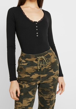 Femme portant une chemise noire à manches longues côtelée avec des boutons et un bord en dentelle, associée à un pantalon cargo camouflage vert et marron.