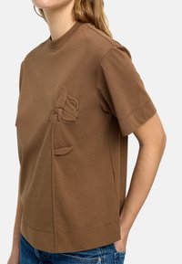 Brązowy t-shirt bawełniany z teksturowanym kwiatowym haftem po boku, okrągłym dekoltem i luźnym krojem. Krótkie rękawy.
