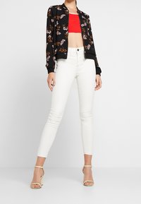 Svart bomberjacka med blommönster, röd kort topp och vita tajta jeans. Modellen bär beiga högklackade sandaler. Släta texturer.