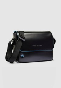 Borsa a tracolla in pelle nera con cuciture e dettagli blu. Presenta una chiusura a patta e una tracolla regolabile, con una texture liscia. Logo del marchio visibile.