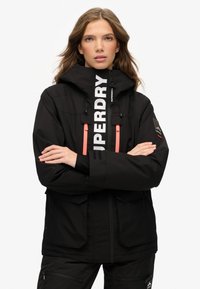 Czarna wodoodporna kurtka z wysokim kołnierzem, zdobiona białym logo, pomarańczowymi zamkami i dwoma przednimi kieszeniami. Gładka tekstura i sportowy design.