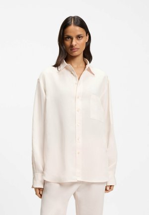 Vrouw met lang donker haar die een losse, lichtroze blouse met borstzak en bijpassende lichte broek draagt, staand tegen een witte achtergrond.