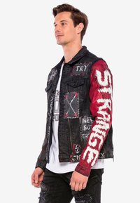 Zwarte spijkerjack met rode mouwen, voorzien van verschillende patches, graffiti-achtige tekst, studs en een ritssluiting aan de voorkant. De buitenkant heeft een licht versleten uitstraling.