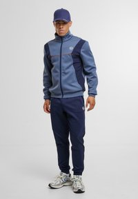 Pánský sportovní komplet v modré a námořnické barvě, obsahující zipovou bundu s barevně dělenými panely a odpovídající tepláky. Zahrnuje logo na hrudi a na kalhotách.